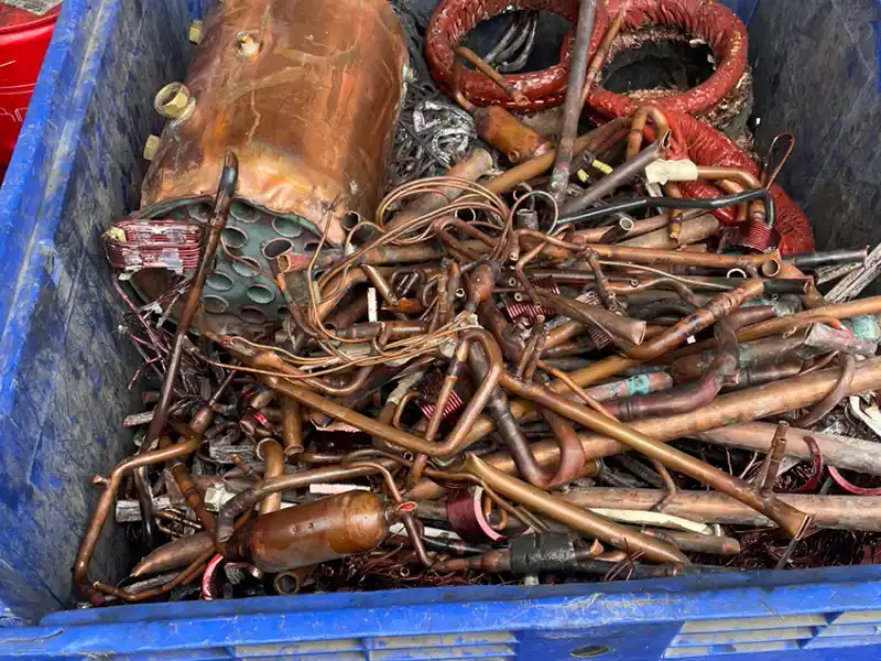 Achat de métaux 91 Un conteneur bleu rempli de divers objets en cuivre usagés, y compris des fils enroulés, des tuyaux et un réservoir cylindrique endommagé - idéal pour Achat de métaux 91. Les pièces métalliques sont enchevêtrées et présentent des signes d'utilisation et d'usure.