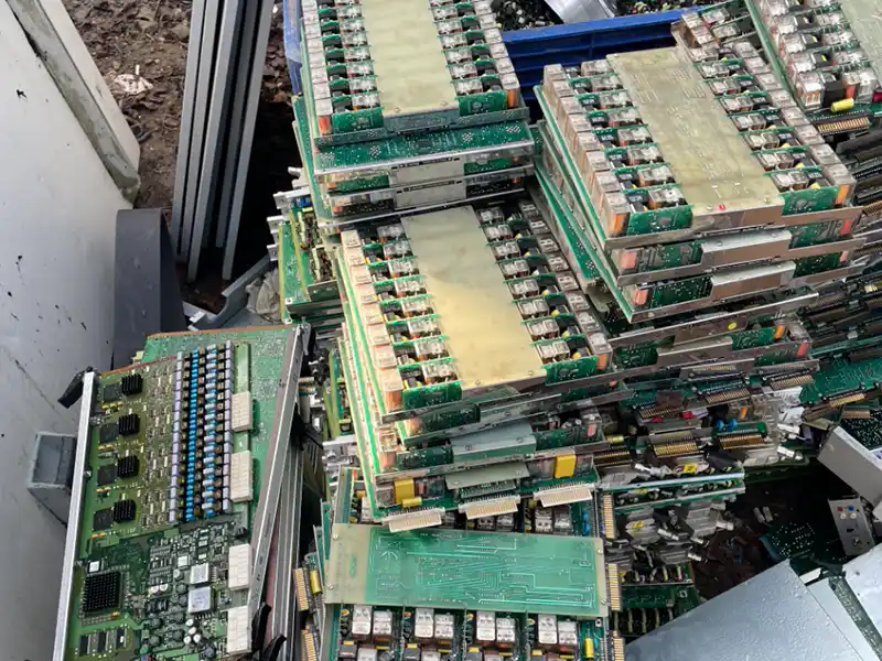 Achat métaux ferreux Un grand tas de circuits imprimés empilés et mis au rebut, avec des puces, des composants et des connecteurs visibles, est situé en plein air, près de métaux, de débris et d'un point de collecte d'Achat métaux ferreux.