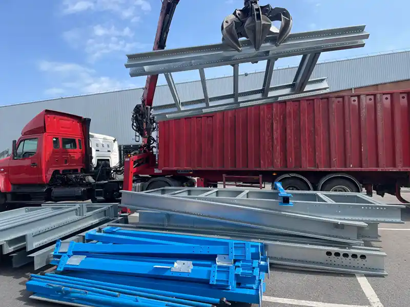Enlèvement des métaux en Essonne Une grue attachée à un camion rouge soulève des poutres métalliques près d'un conteneur maritime rouge, dans le cadre de l'enlèvement des métaux en Essonne, avec des composants métalliques bleus et gris empilés sur le sol dans une zone industrielle.