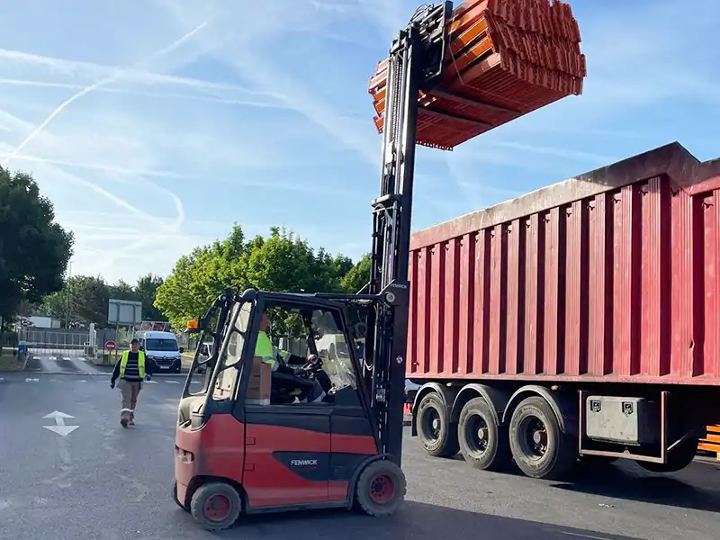 Entreprise Ferrailleur Essonne - Baud Un chariot élévateur à fourche soulève une pile de barrières orange au-dessus d'un grand conteneur rouge dans un parking, tandis qu'une personne portant un gilet de sécurité d'une Entreprise de Ferrailleur en Essonne marche à proximité. Des arbres et une camionnette blanche se trouvent à l'arrière-plan.