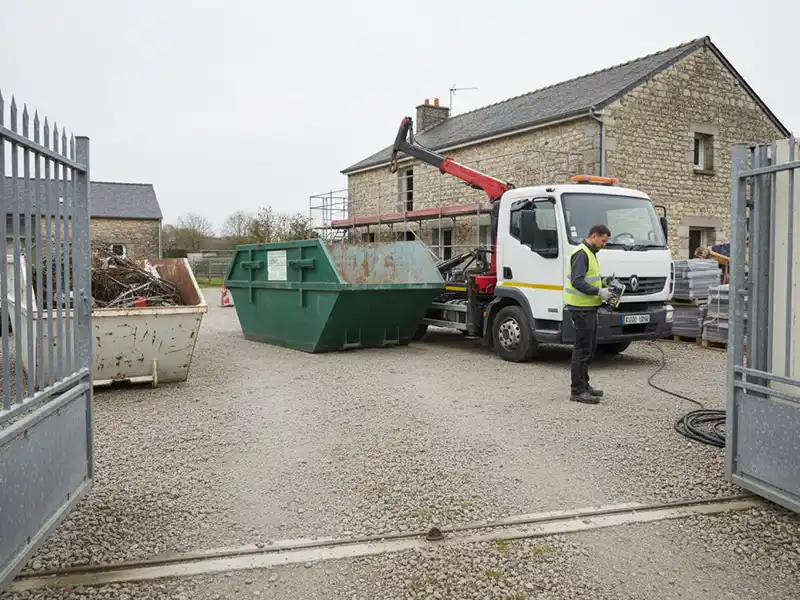 Location de benne en Essonne Un ouvrier se tient à côté d'un camion blanc avec une grue, déchargeant une grande benne verte dans une cour en gravier près d'un bâtiment en pierre ; une autre benne remplie se trouve à proximité, illustrant la location de benne en Essonne.